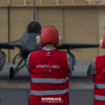 Tres armeros de línea de la Fuerza Aérea de Chile con chalecos rojos observan un caza F-16 frente al hangar.