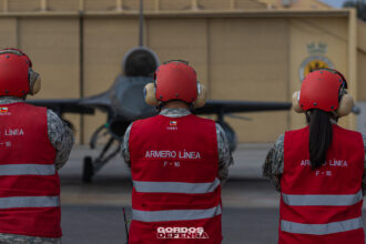 Tres armeros de línea de la Fuerza Aérea de Chile con chalecos rojos observan un caza F-16 frente al hangar.