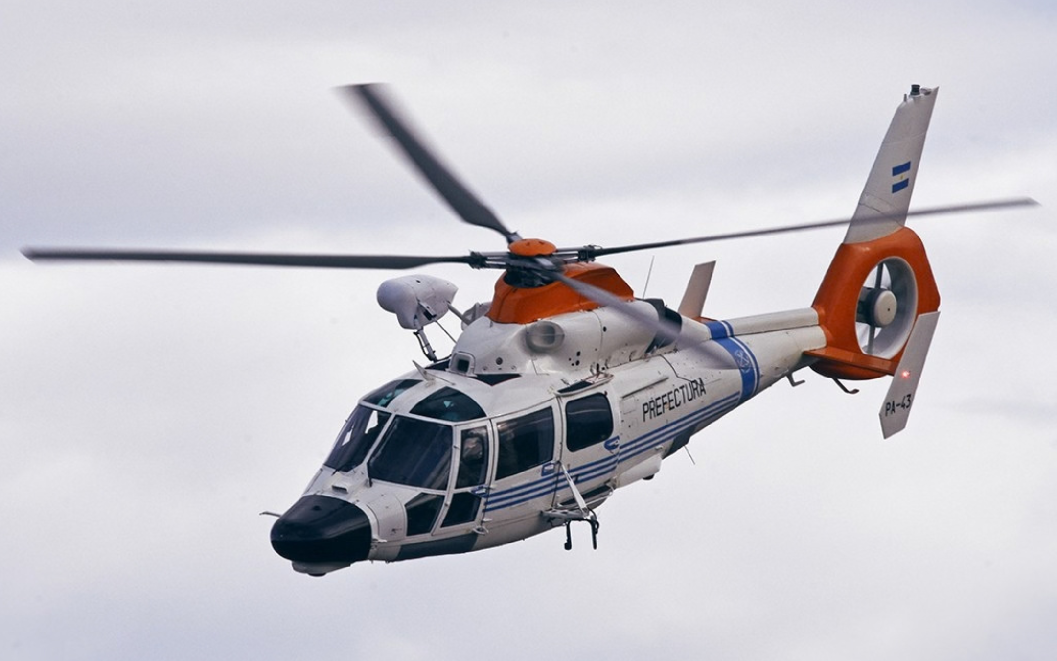 Helicóptero AS365 Dauphin matrícula PA-43 de la aviación de Prefectura Naval en vuelo, con torreta electroóptica sobre la cabina y fenestrón naranja de alta visibilidad.
