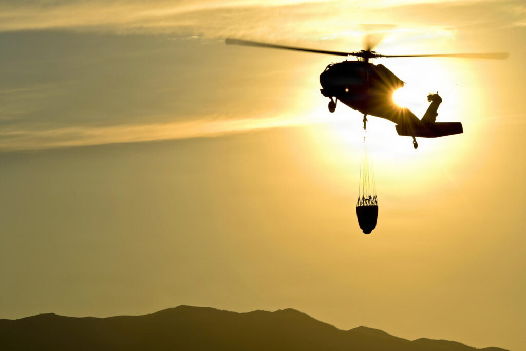 Helicóptero Black Hawk transportando un balde Bambi Bucket suspendido por cable al atardecer sobre terreno montañoso