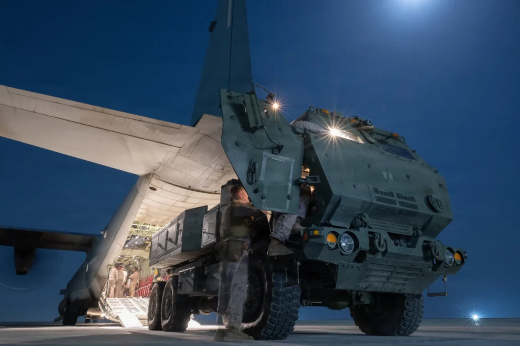 Lanzador M142 HIMARS junto a la rampa trasera abierta de un avión de transporte C-130, de noche, con personal militar a su alrededor
