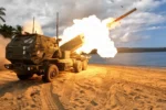 Lanzador de cohetes M142 HIMARS disparando un cohete desde una playa, con palmeras y el mar de fondo