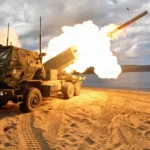 Lanzador de cohetes M142 HIMARS disparando un cohete desde una playa, con palmeras y el mar de fondo
