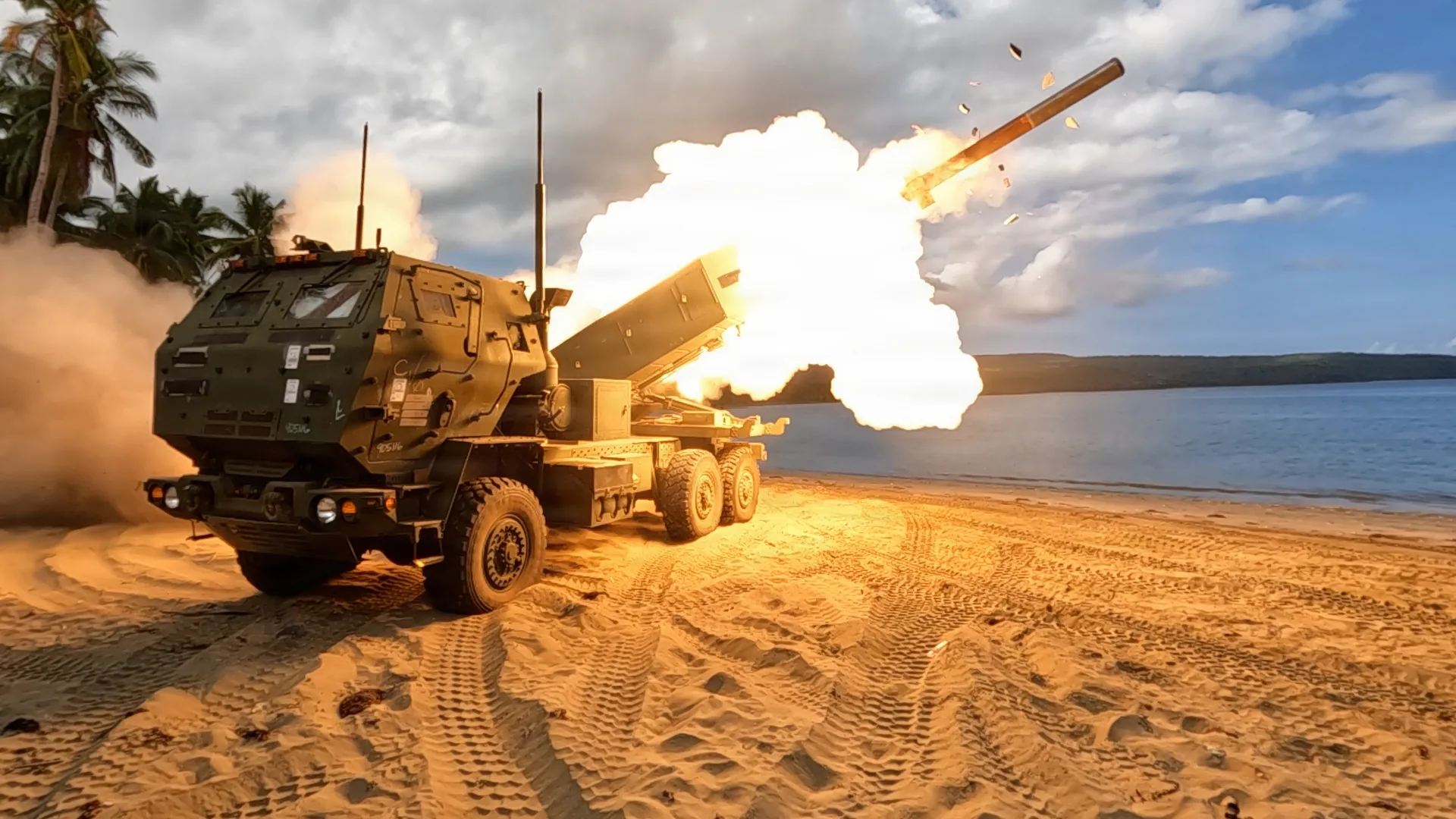 Lanzador de cohetes M142 HIMARS disparando un cohete desde una playa, con palmeras y el mar de fondo