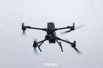 Dron cuadricóptero negro con una cámara multisensor montada debajo, visto desde abajo en vuelo contra el cielo nublado