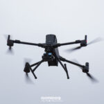 Dron cuadricóptero negro con una cámara multisensor montada debajo, visto desde abajo en vuelo contra el cielo nublado