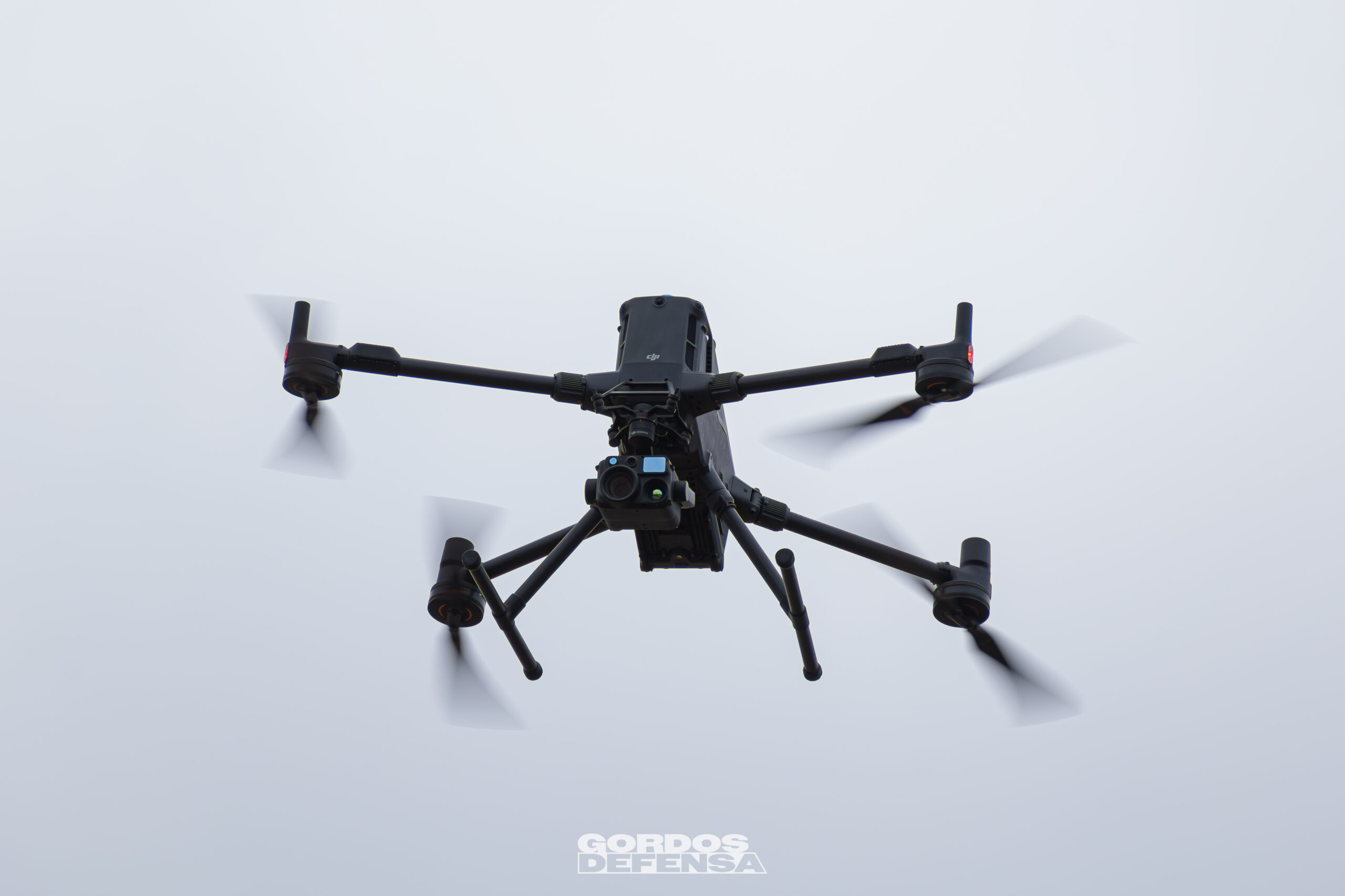 Dron cuadricóptero negro con una cámara multisensor montada debajo, visto desde abajo en vuelo contra el cielo nublado