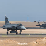 Caza Gripen gris con el número 4106 en la deriva rodando por una pista en zona desértica, con un helicóptero Black Hawk detrás