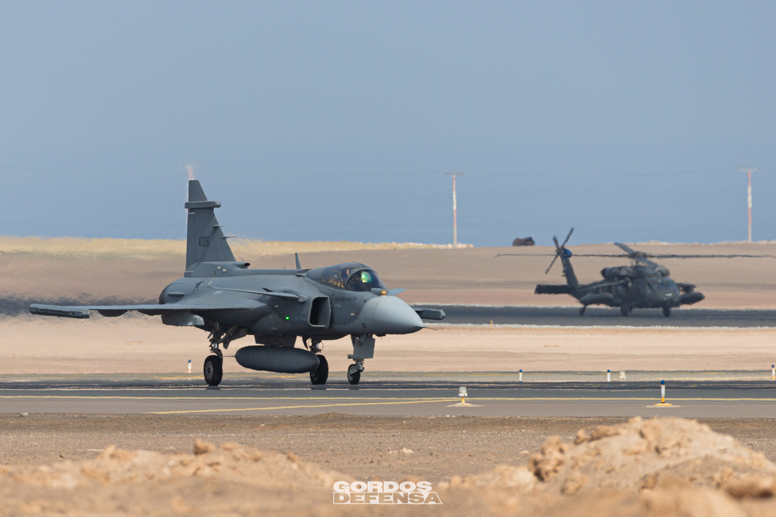Caza Gripen gris con el número 4106 en la deriva rodando por una pista en zona desértica, con un helicóptero Black Hawk detrás